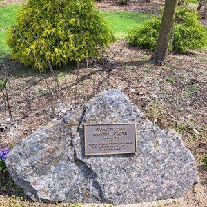 Memorial plaque on a large rock in front of green trees and purple flowers: text in caption below