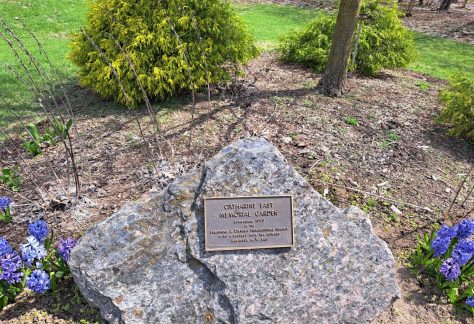 Memorial plaque on a large rock in front of green trees and purple flowers: text in caption below